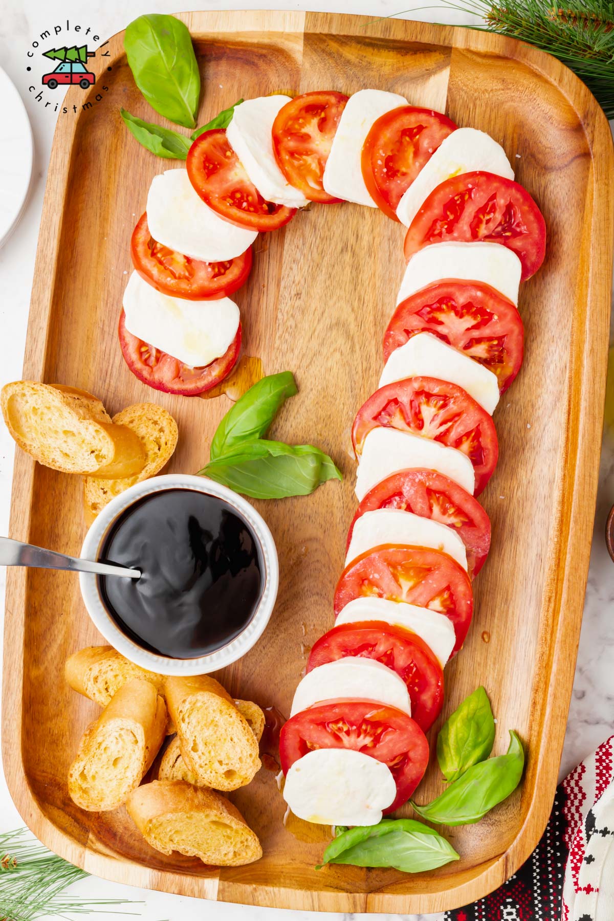 Caprese salad ingredients shaped into a candy cane with basil sprinkled on the platter and a bowl of balsamic vinaigrette and toasted baguette slices.