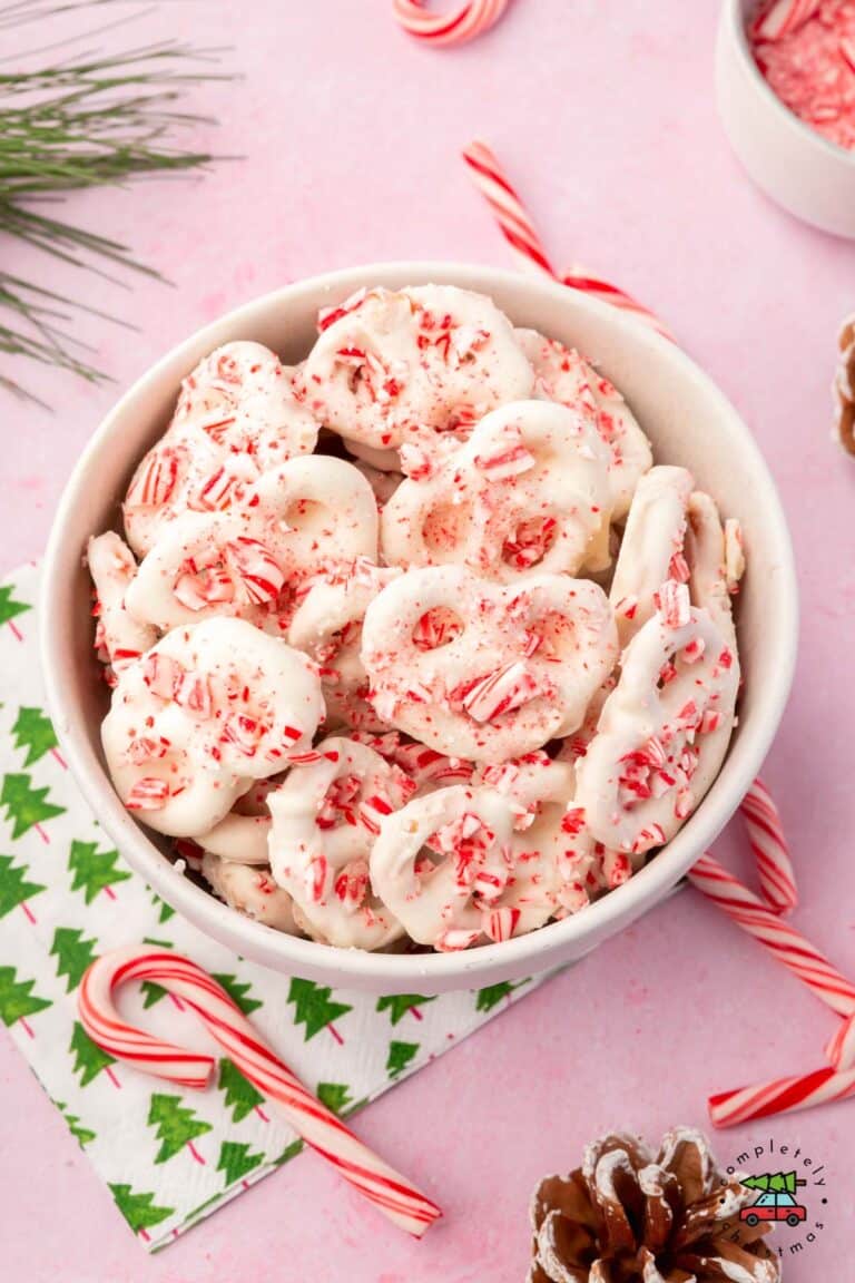 A bowl of white chocolate covered pretzels topped with crushed up candy canes with a christmas tree napkin underneath it on a pink table.