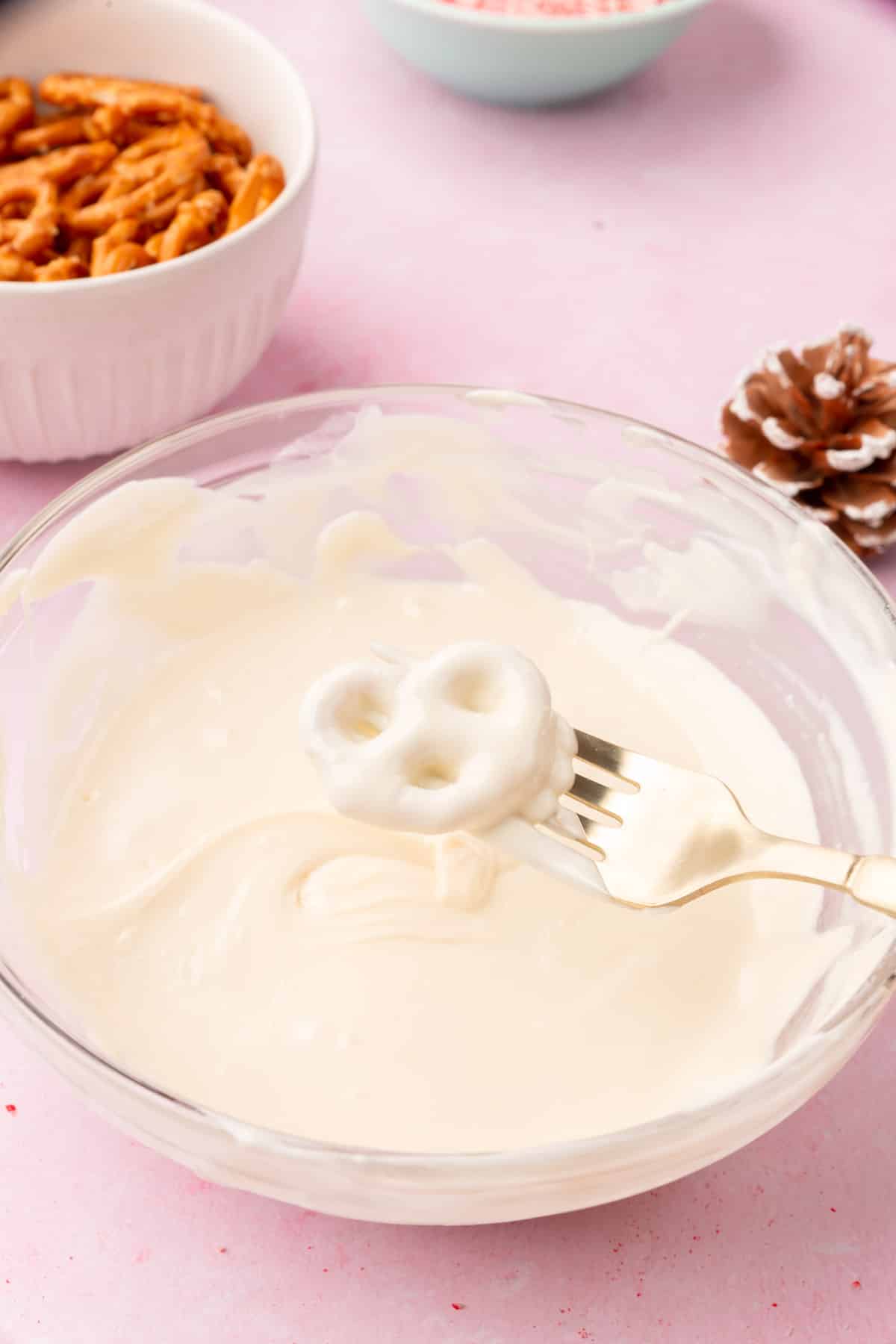 A bowl of melted white chocolate with a fork holding a dipped pretzel over the bowl.