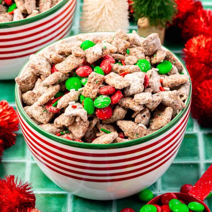 A bowl of Christmas puppy chow topped with M&Ms on a green tile table with Christmas decor surrounding it.