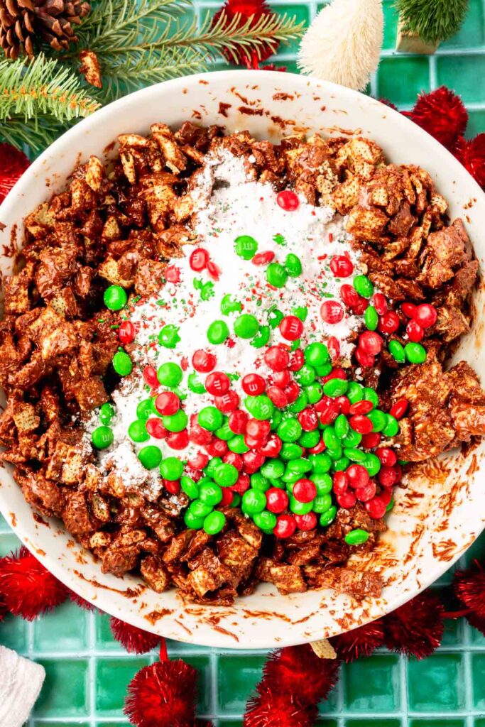 A large bowl of rice chex cereal covered with melted chocolate and topped with powdered sugar, holiday sprinkles and red and green M&Ms before mixing.