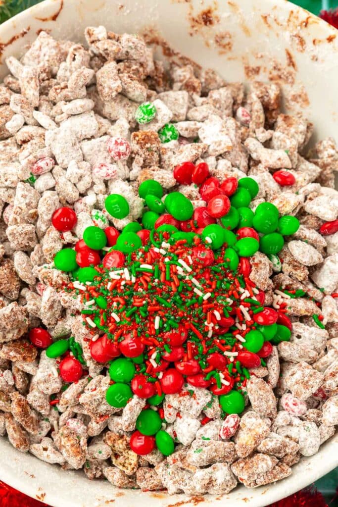 A large bowl of puppy chow that has been topped with red and green M&Ms and holiday sprinkles before mixing.