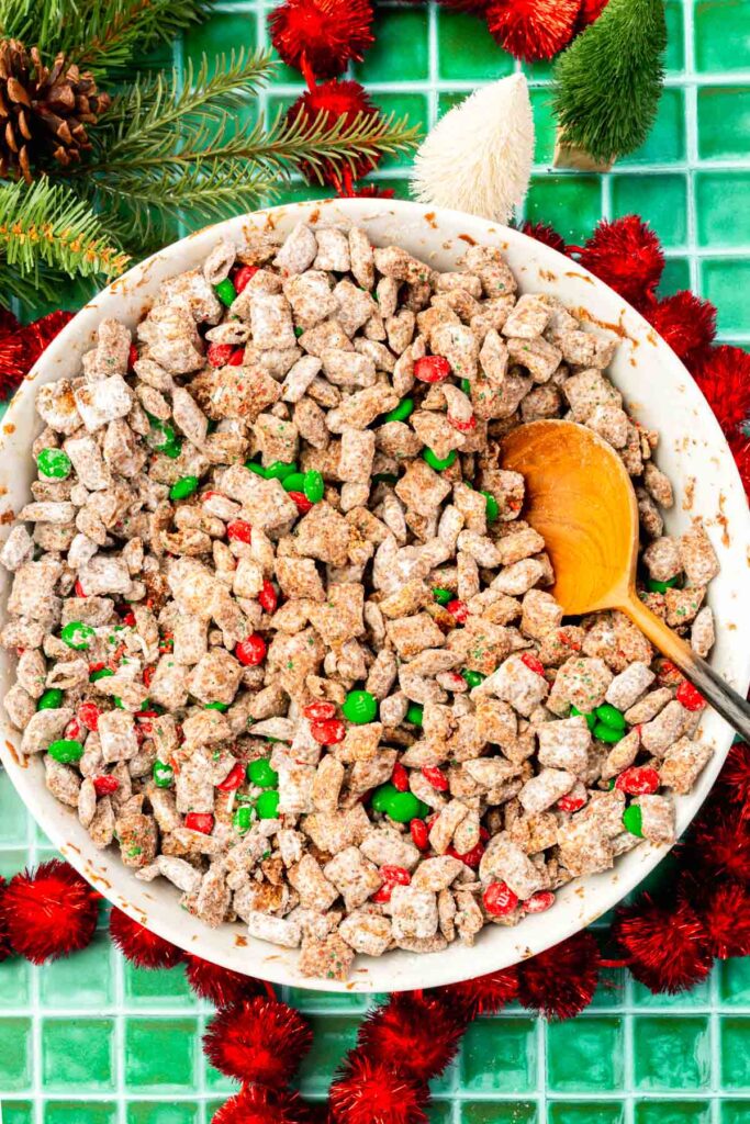 A large mixing bowl of Christmas puppy show topped with M&Ms with a wooden serving spoon in it.
