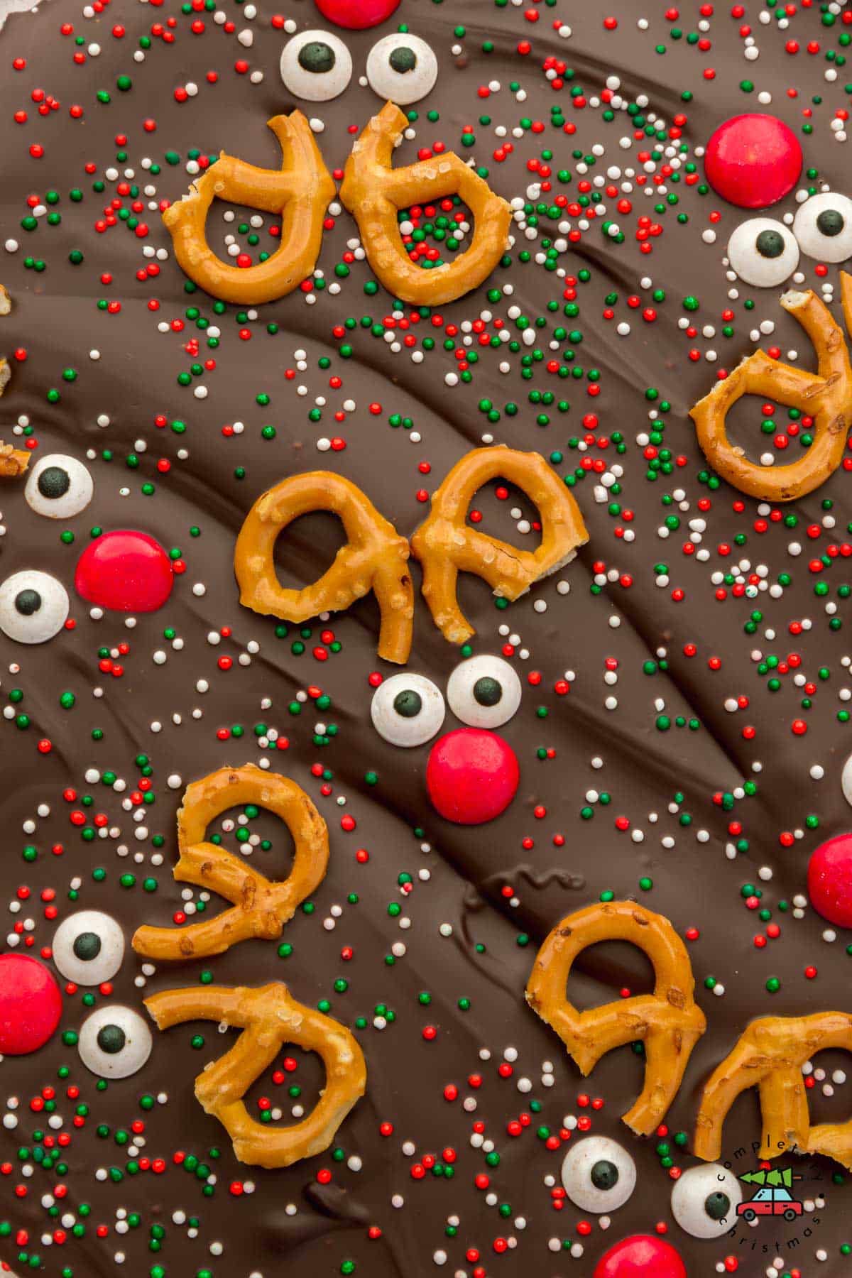 A closeup of reindeer chocolate bark topped with pretzels, red M&M, candy eye sprinkles, and Christmas colored sprinkles before slicing.