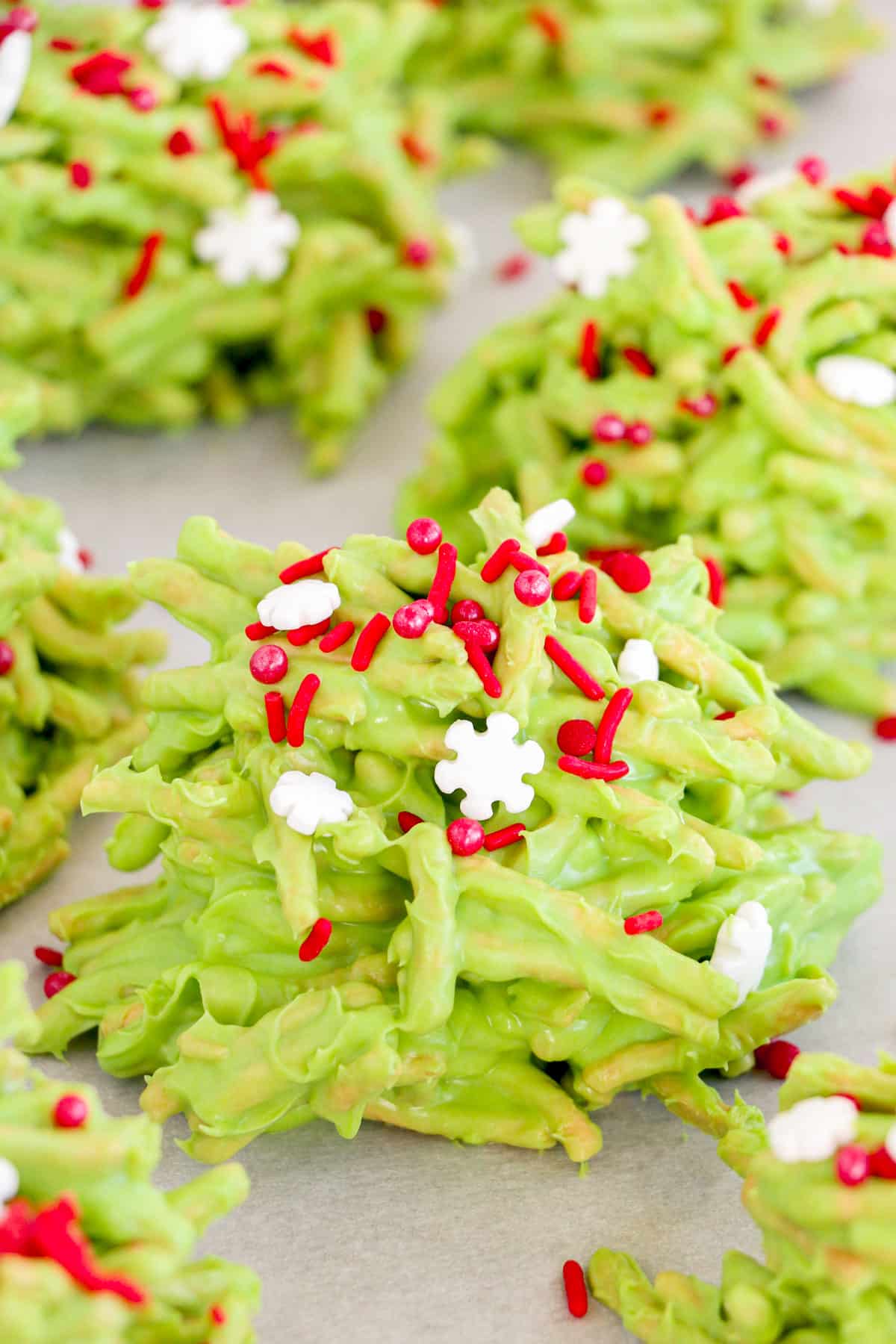 A green chow mein noodle cookie topped with red and snowflake sprinkles on a piece of parchment paper.