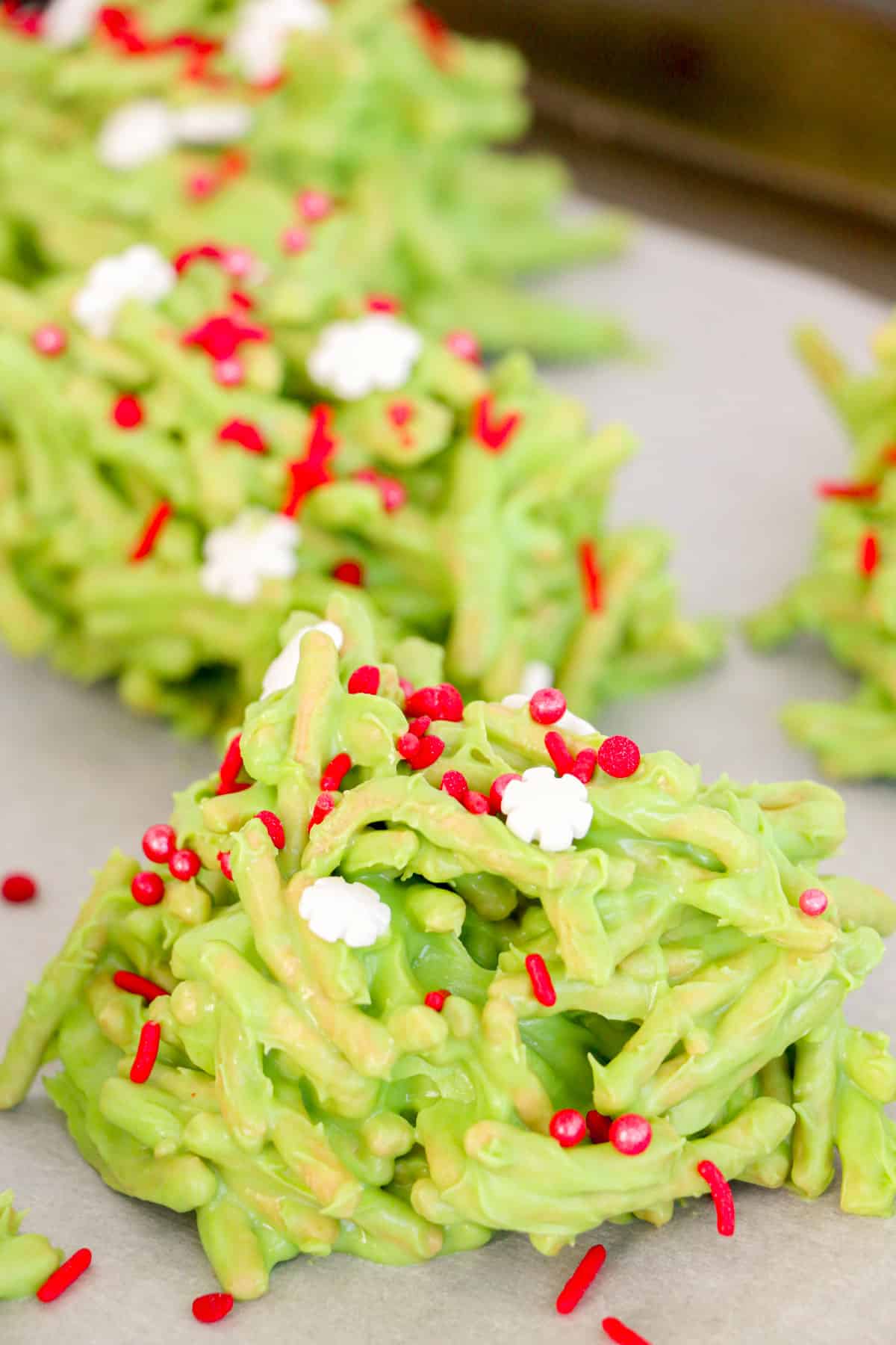 Multiple green haystack cookies topped with Christmas sprinkles on a parchment lined baking sheet.