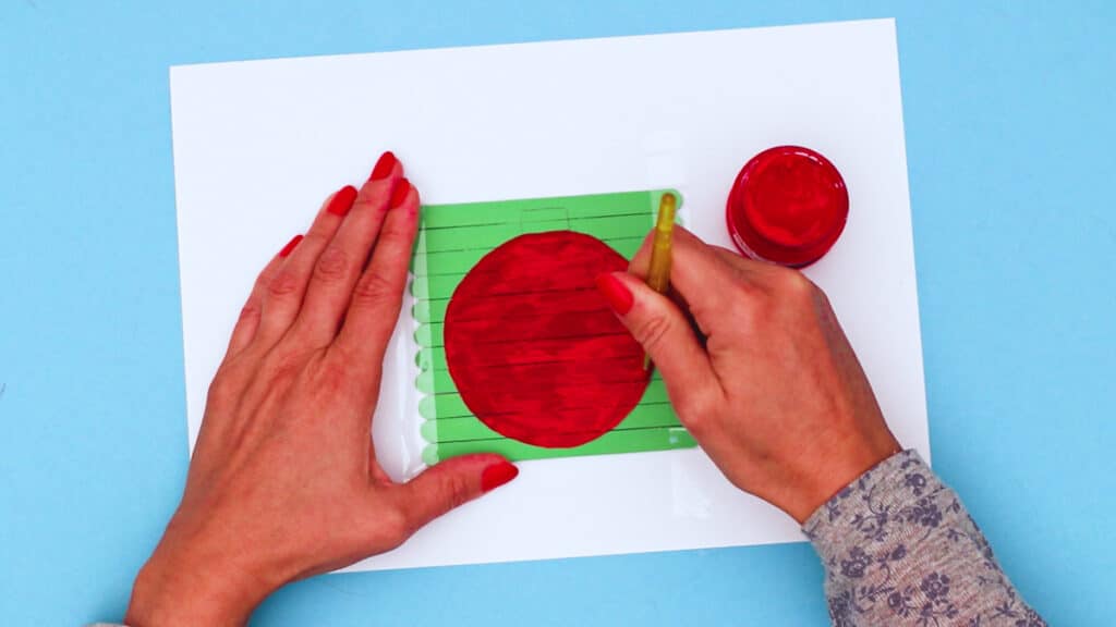 Two hands painting an ornament red on top of green popsicle sticks.