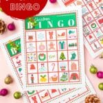 A printable Christmas Bingo card on a pink table with mini ornaments and pine cones on the surface.