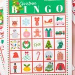 A printable Christmas Bingo card on a pink table with mini ornaments and pine cones on the surface.