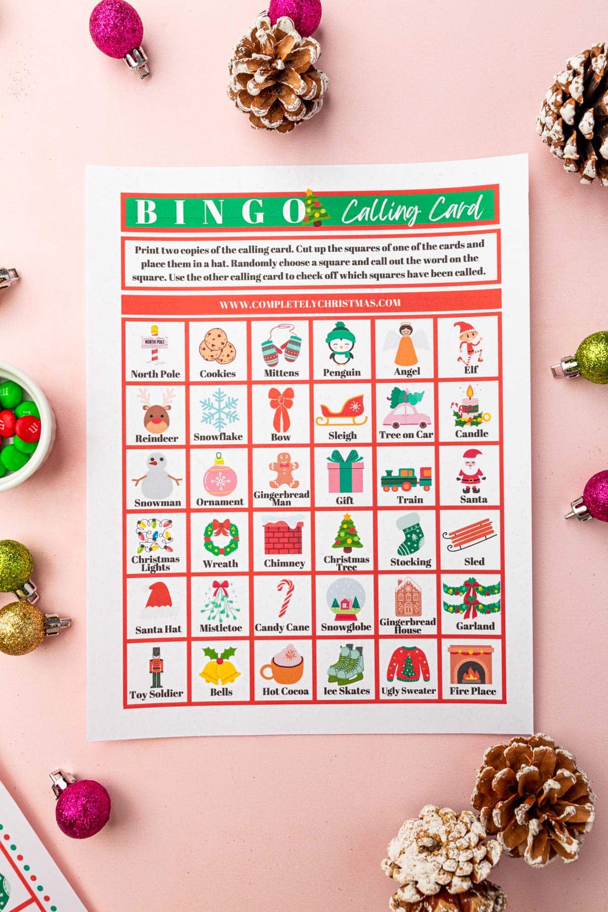 A printable Christmas Bingo calling card on a pink table with mini ornaments and pine cones on the surface.