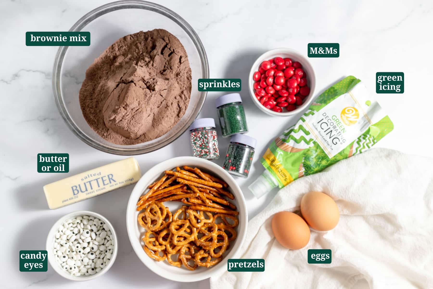 Ingredients in small bowls on a table for making Christmas brownies, including brownie mix, sprinkles, M&Ms, green icing, pretzels, eggs, butter, and candy eyes with text overlays over each ingredient.