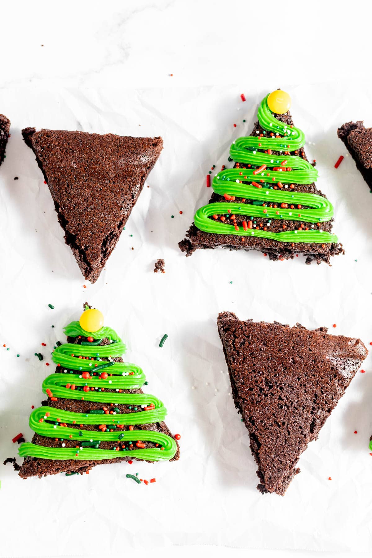 Four Christmas brownies in the shape of triangles and decorated like Christmas trees with green frosting, M&Ms and Christmas sprinkles.