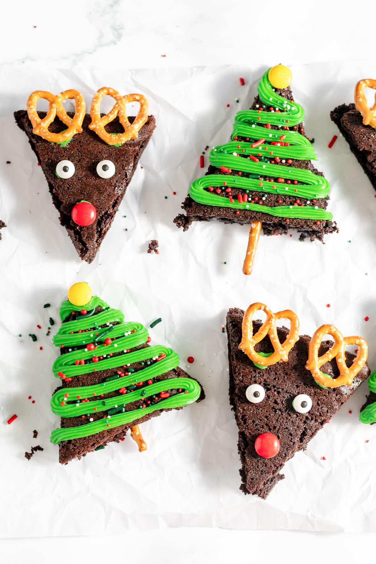 Four Christmas brownies in the shape of triangles and decorated like Christmas trees and reindeer with green frosting, candy eyes, M&Ms and pretzels.