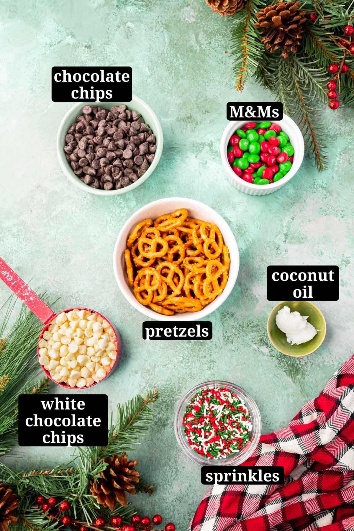 Small bowls of ingredients on a green table to make Christmas pretzels, including semi-sweet chocolate chips, red and green M&Ms, white chocolate chips, pretzels, coconut oil, and Christmas sprinkles with text overlays over each ingredient.