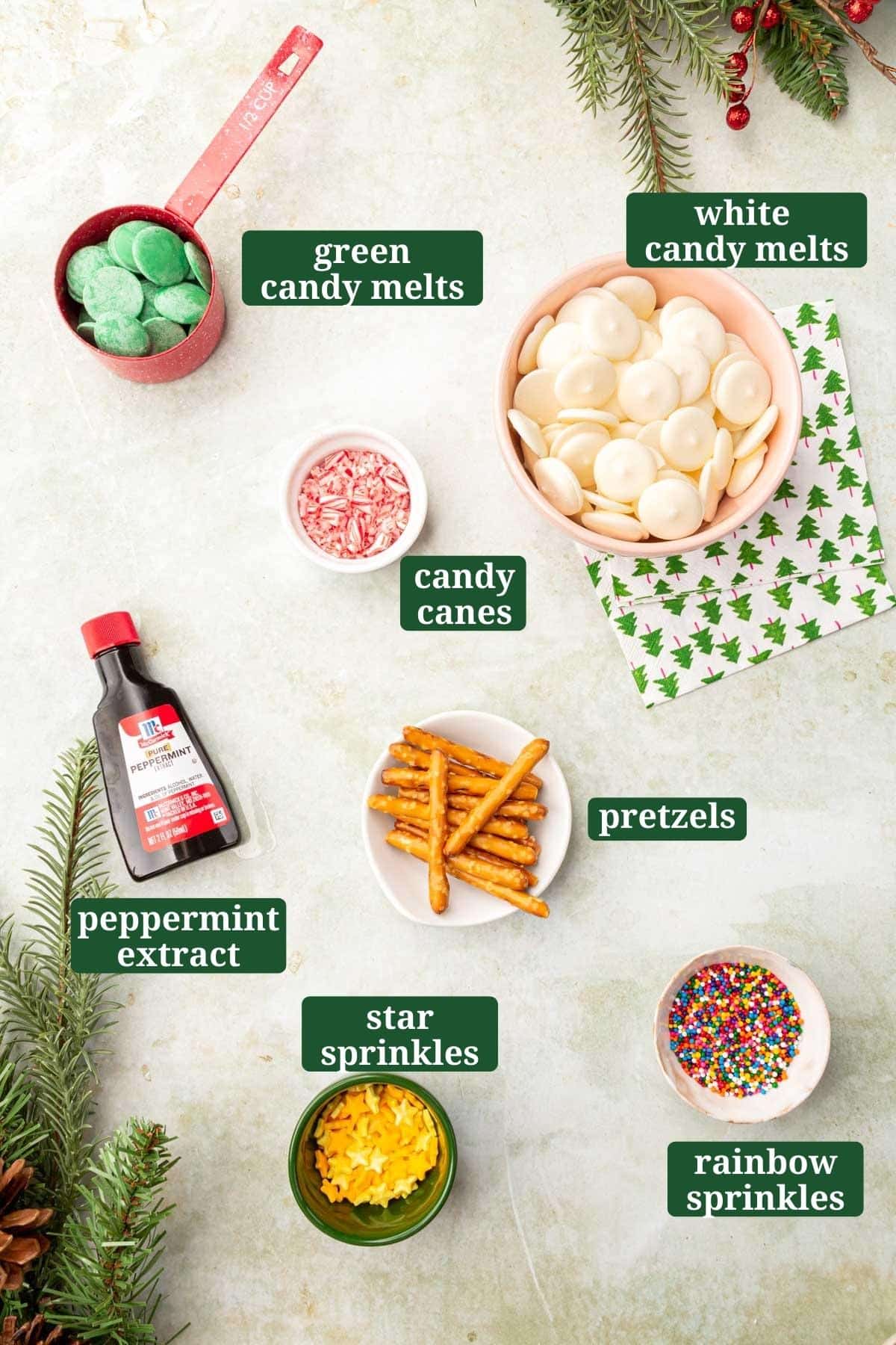Small bowls of ingredients on a green table to make Christmas tree white peppermint candy bark, including candy canes, pretzels, peppermint extract, sprinkles with text overlays over each ingredient.