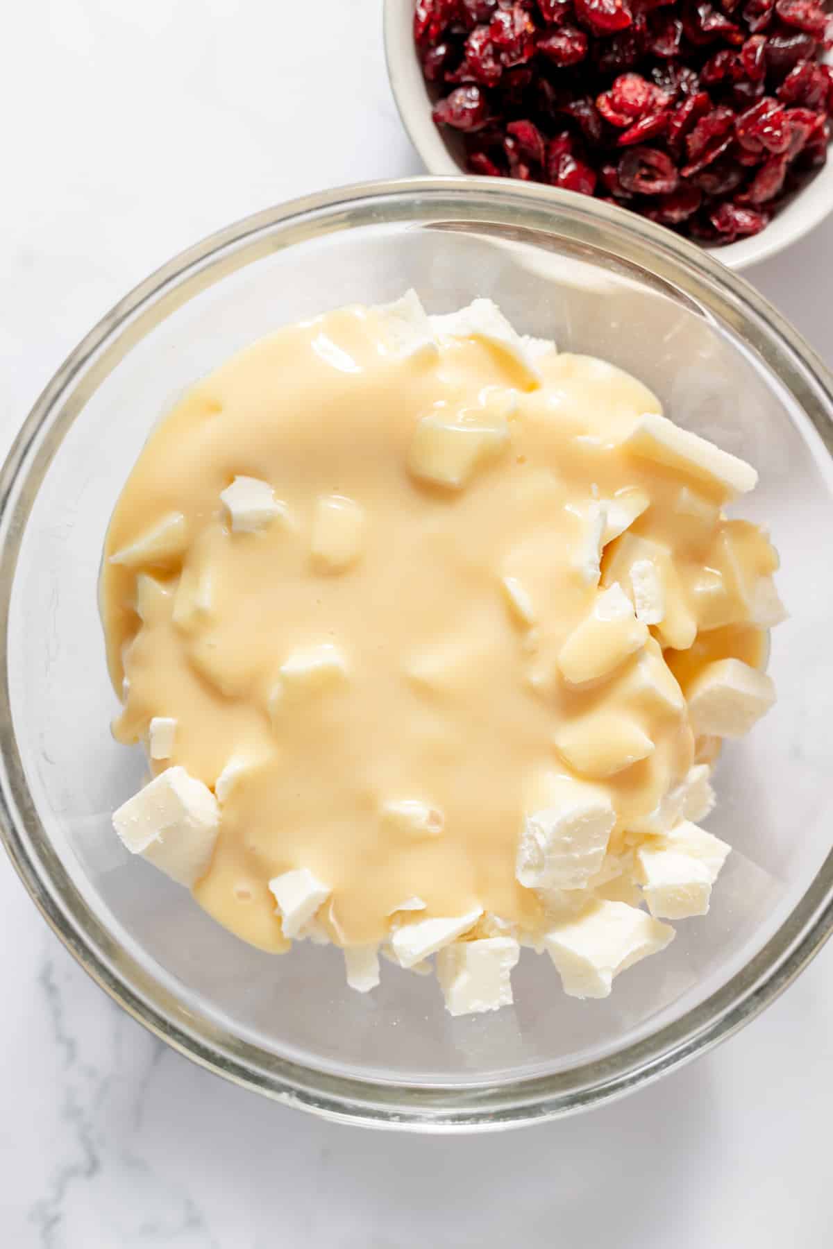 A glass mixing bowl with chopped white chocolate and sweetened condensed milk with a bowl of dried cranberries to the side.