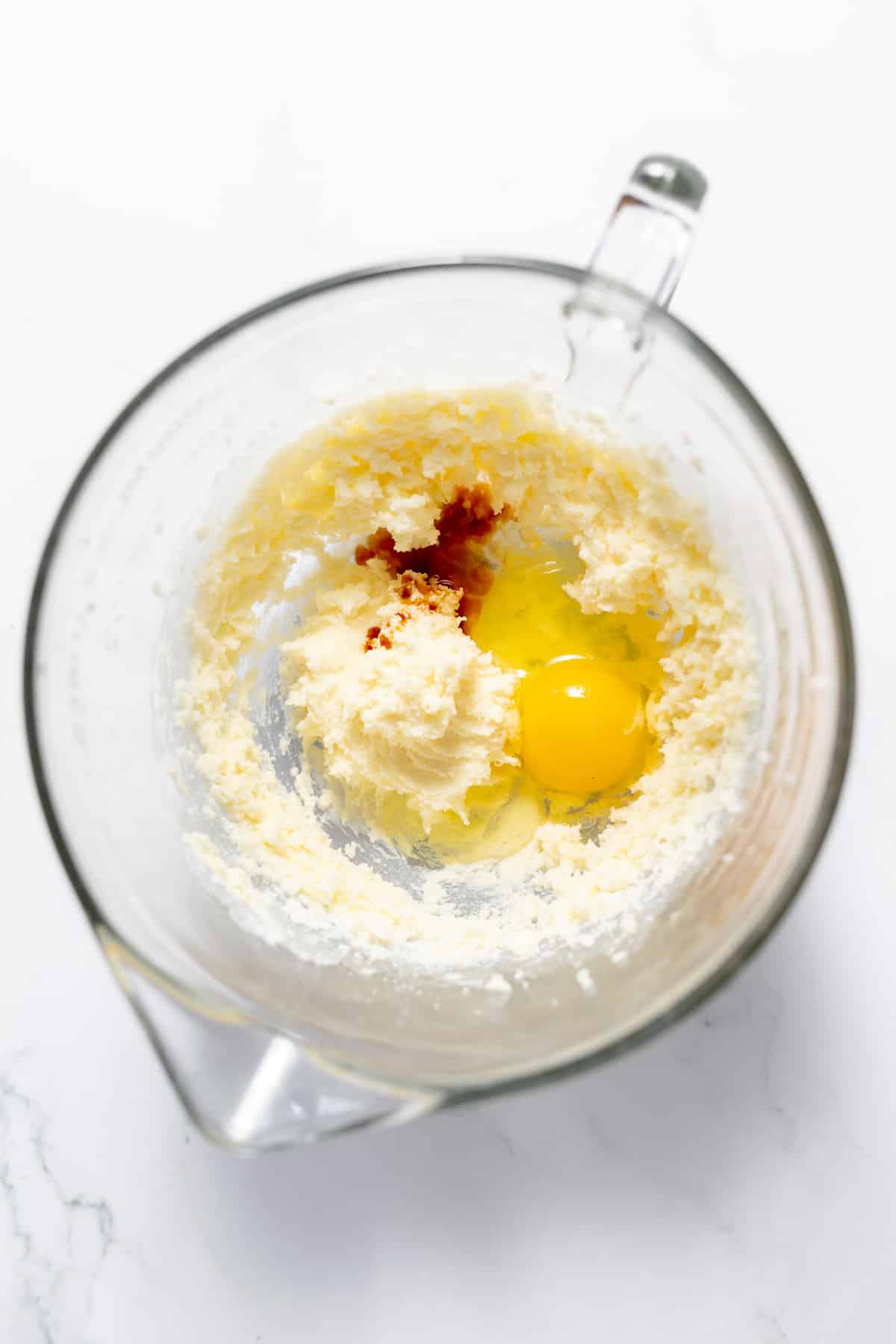A glass mixing bowl with creamed butter and sugar topped with an egg and vanilla extract.