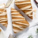 An overhead view of three gingerbread scones with a drizzled glaze on a white table with forks, greenery, and a knife.