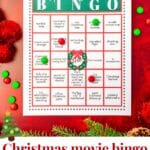 A printable Hallmark Christmas movie bingo card on a red table with green and red M&Ms, red pom pom garland, and greenery on the surface.