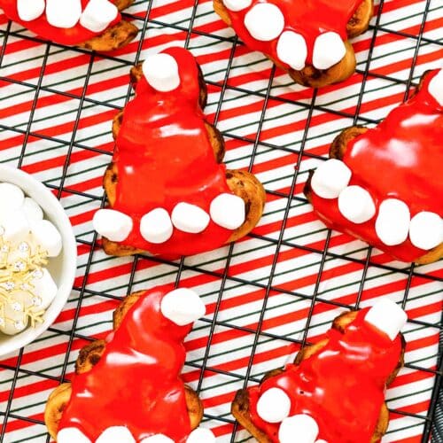 Cinnamon rolls decorated to look like Santa hats with red icing and mini marshmallows on a wire rack with a red striped towel.
