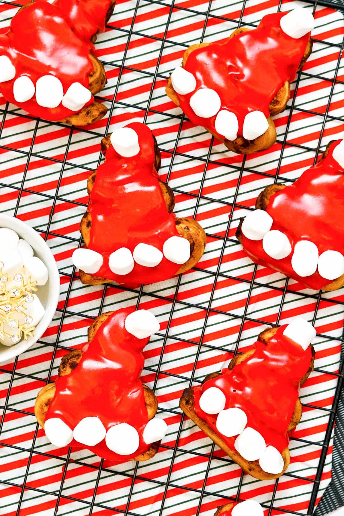 Cinnamon rolls decorated to look like Santa hats with red icing and mini marshmallows on a wire rack with a red striped towel.