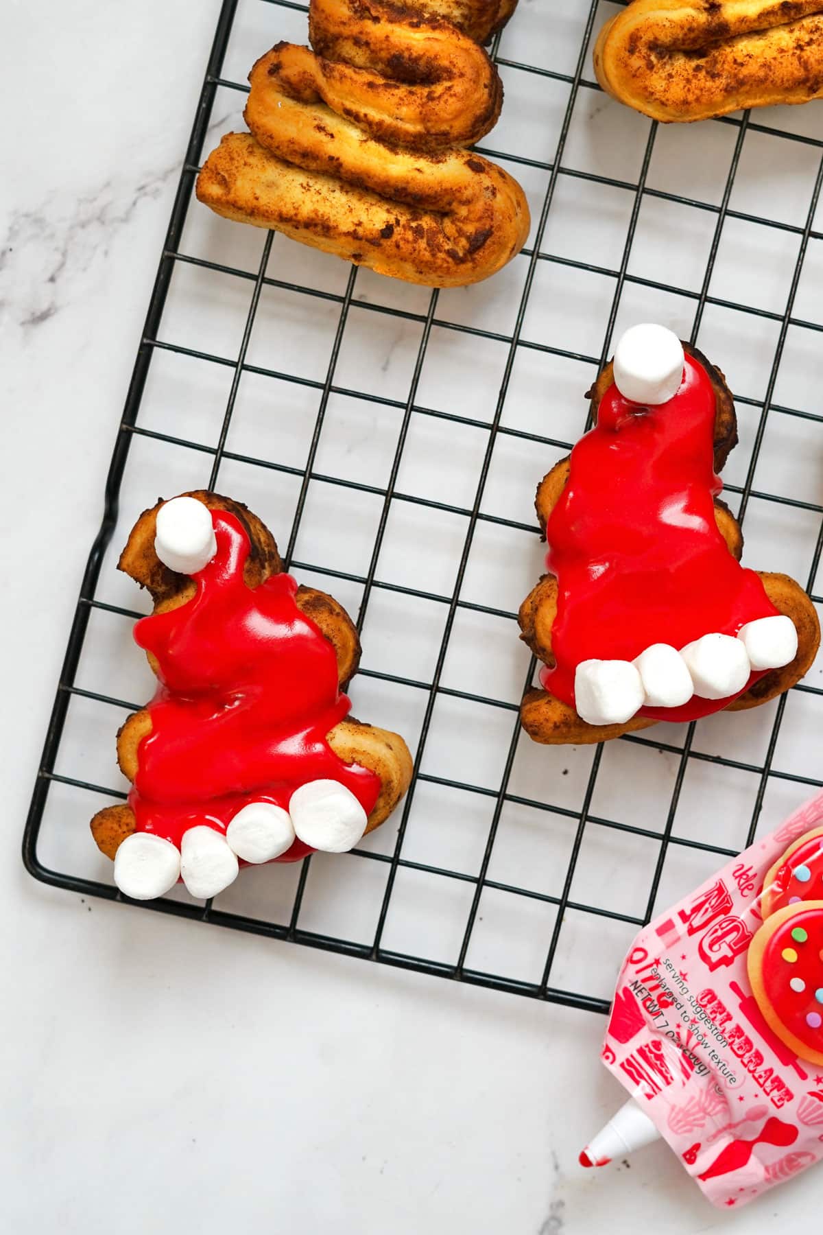 Cinnamon rolls in the shape of a Santa hat on a cooling rack with a few decorated with red icing and mini marshmallows.