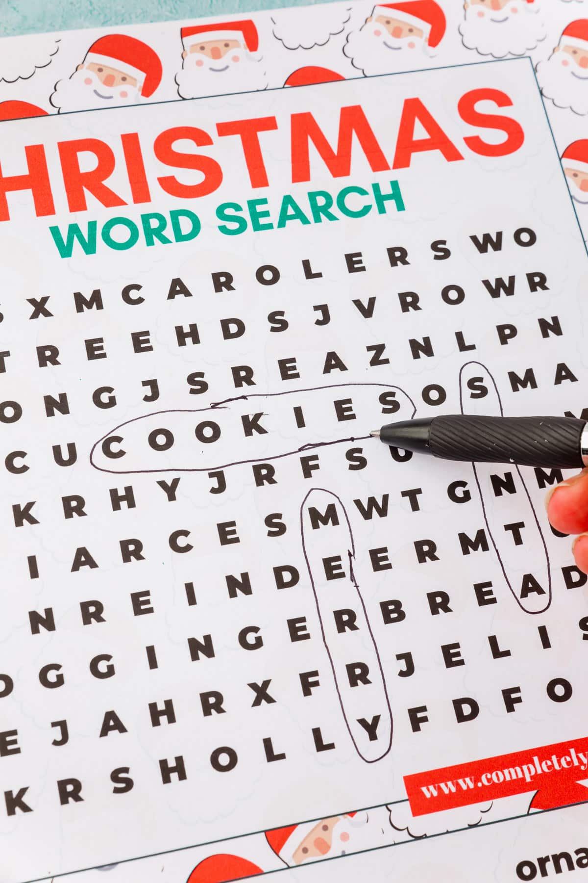 A hand circling the word "cookies" on a Christmas word search printable.