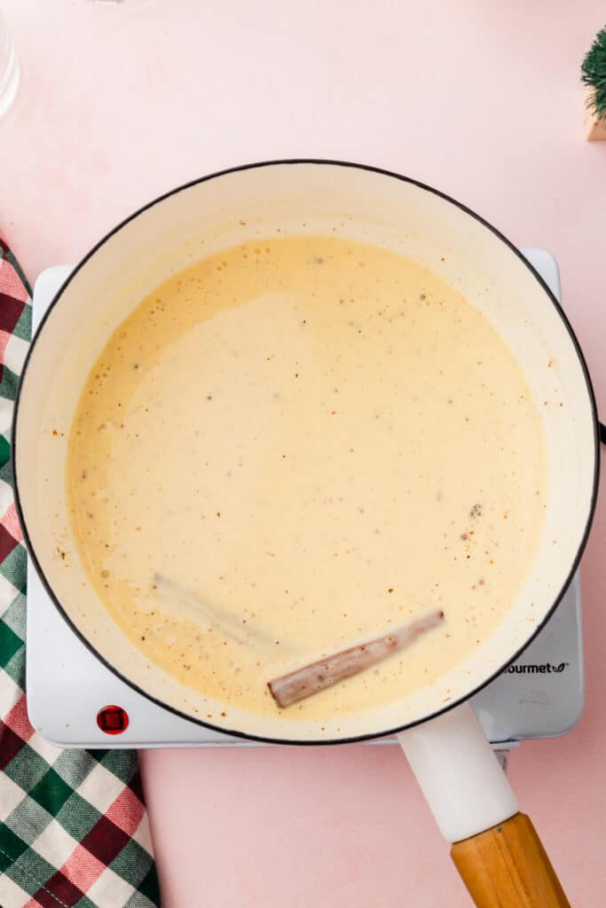A saucepan filled with eggnog mixed together with ground nutmeg and two cinnamon sticks.