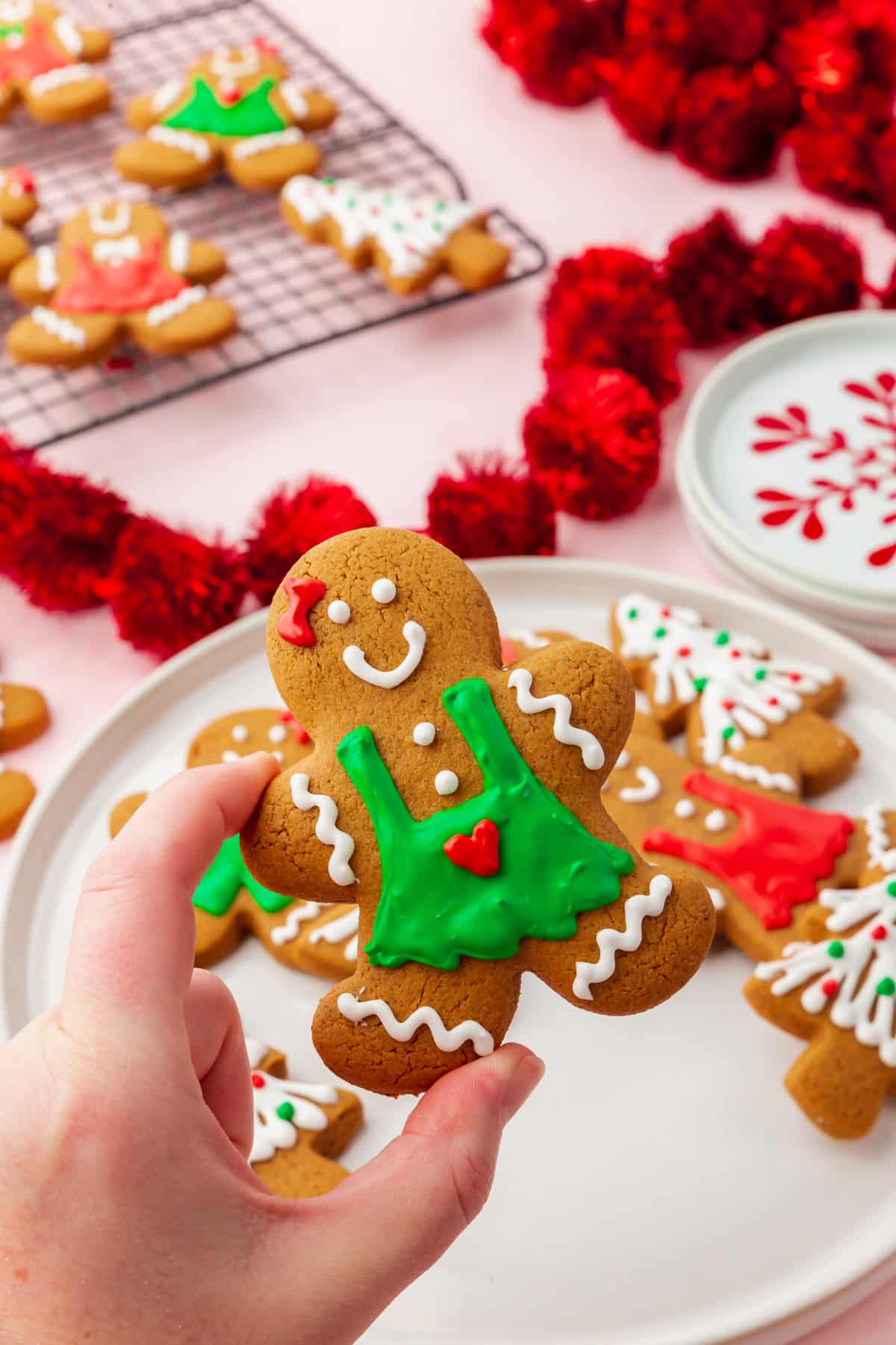 A hand holding a gingerbread person cookie decorated with a green royal icing dress and white and red decorations.