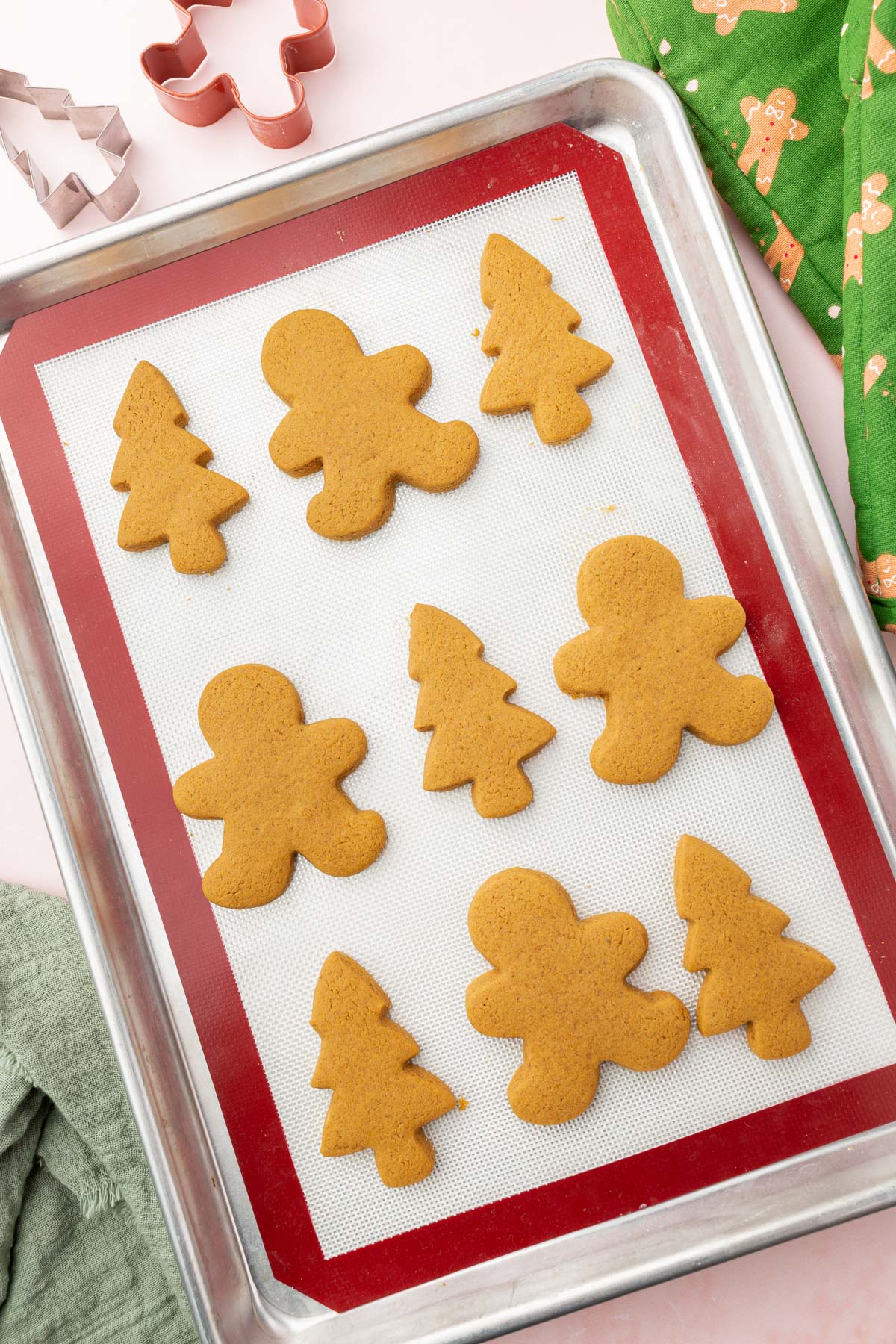 A baking sheet lined with a silicone baking mat and topped with gingerbread cookies cut out into Christmas tree and gingerbread men shapes after baking in the oven.