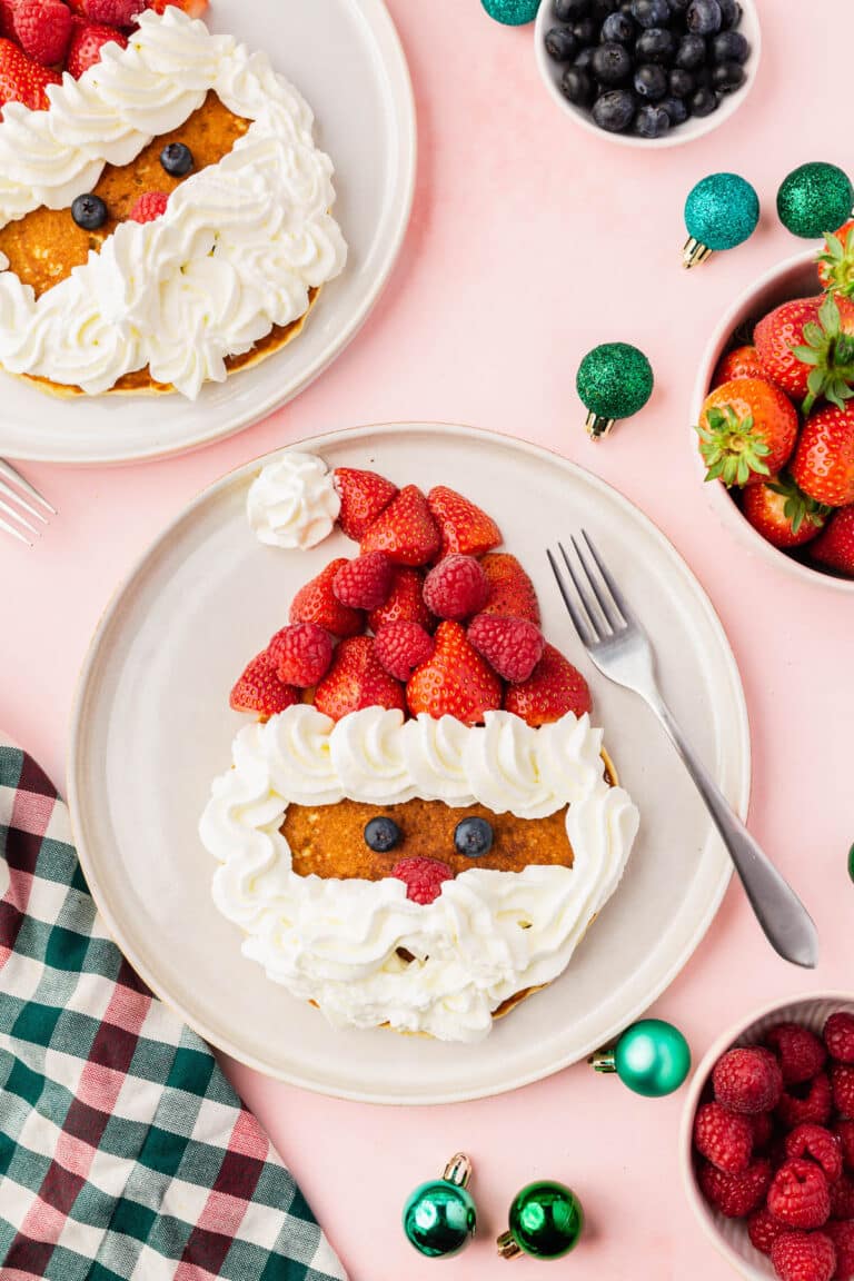 A large pancake decorated with strawberries, raspberries, and whipped cream to look like Santa Claus with bowls or berries and ornaments around it.
