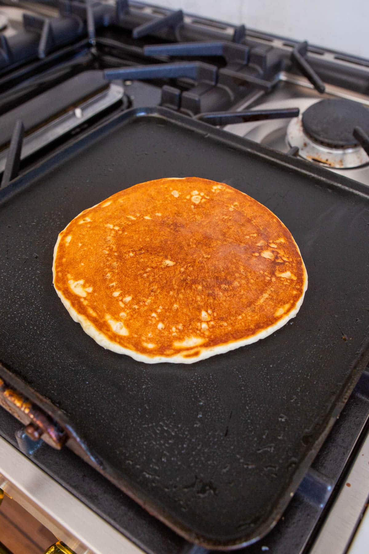 A large pancake cooking on a square non-stick griddle pan on a stove-top.