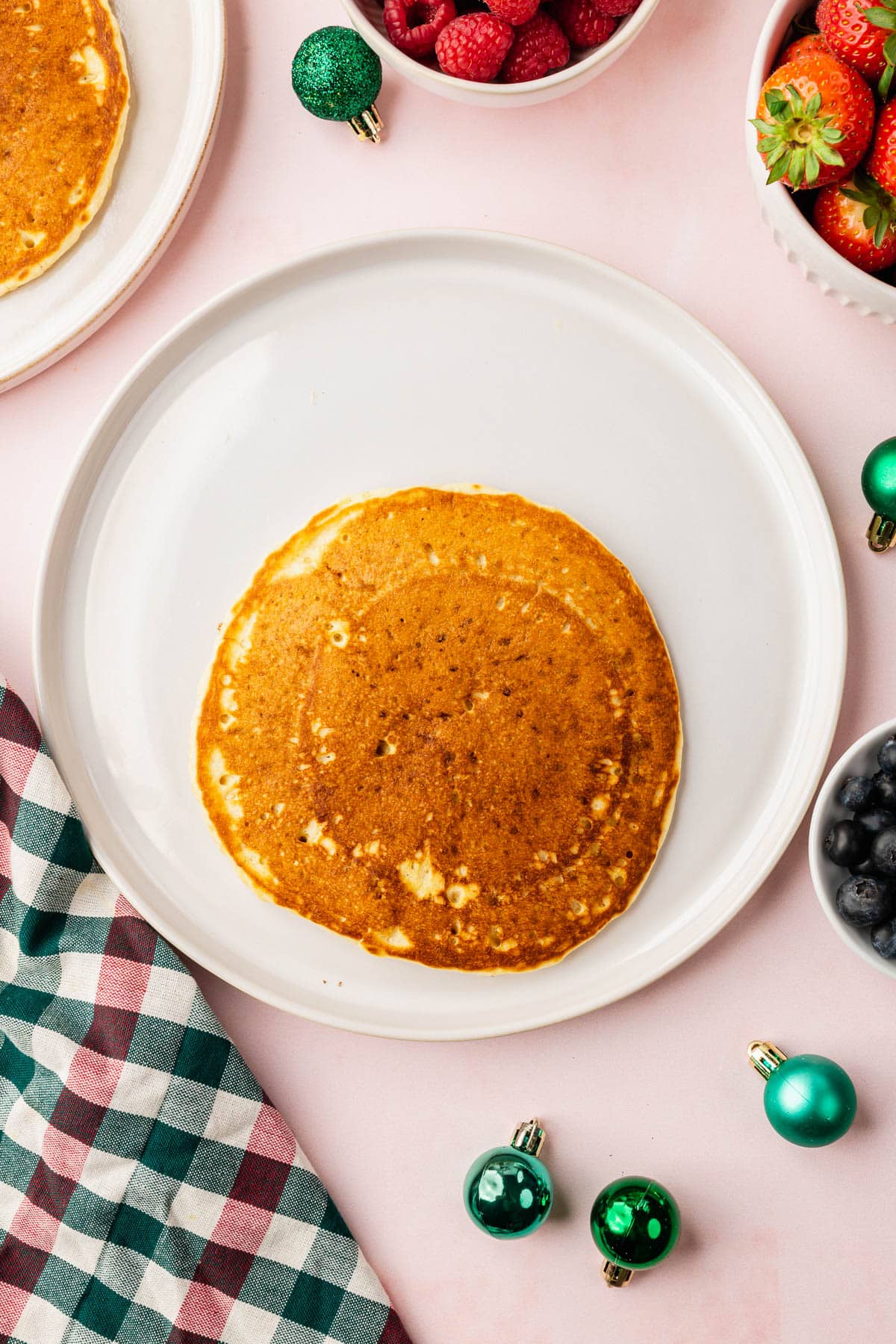 A single large pancake on a plate surrounded by bowls of berries and ornaments.