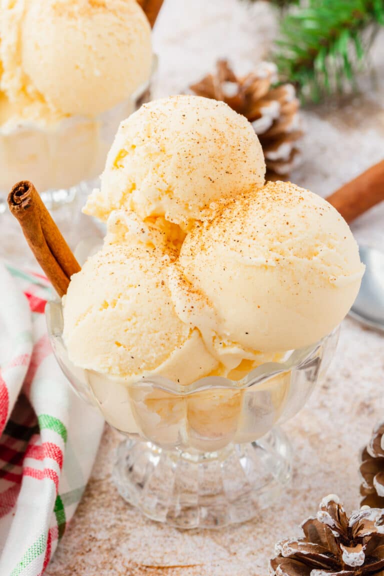 A glass dish filled with scoops of eggnog ice cream sprinkled with ground nutmeg and a cinnamon stick inserted into the dish.