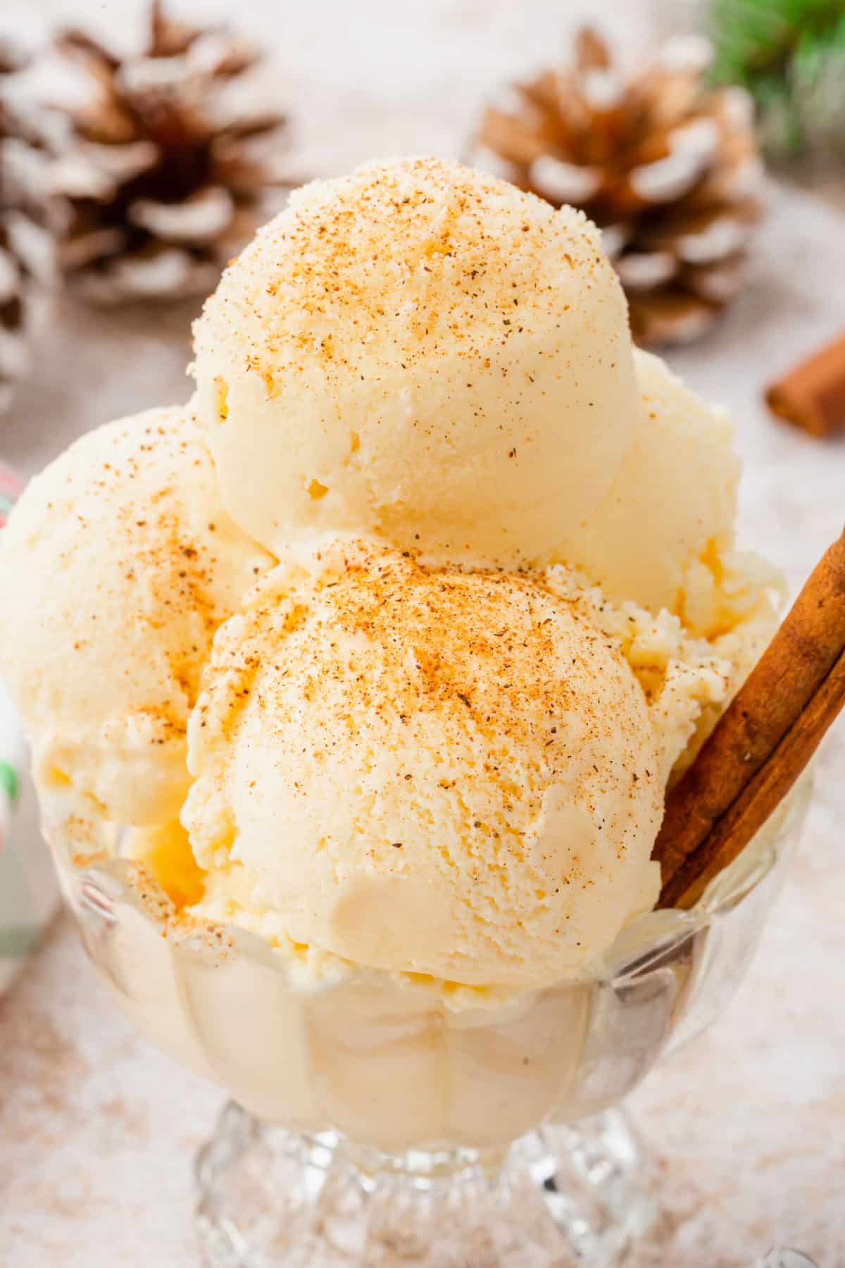 A glass dish filled with scoops of eggnog ice cream sprinkled with ground nutmeg and a cinnamon stick inserted into the dish.