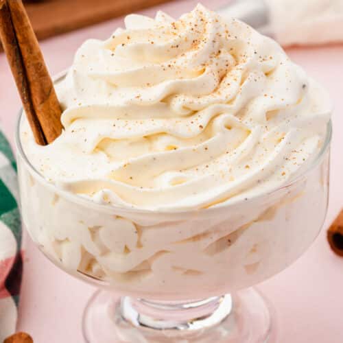 A glass dish filled with piped eggnog whipped cream topped with a sprinkle of nutmeg and a cinnamon stick.