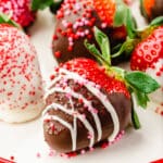 A closeup of a strawberry decorated in dark chocolate, a drizzle of white chocolate, and red and pink sprinkles for Valentine's Day with more strawberries behind it.