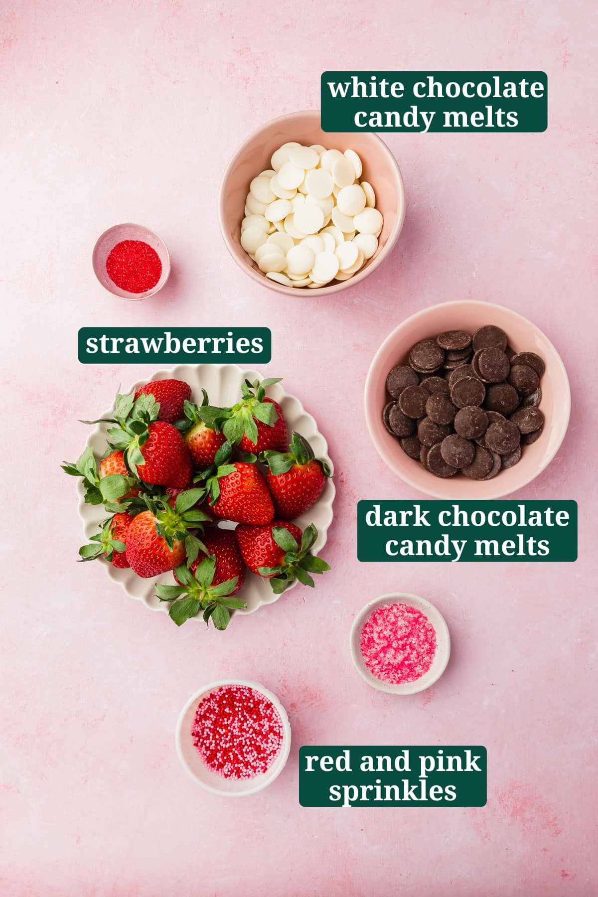 An overhead view of small bowls of ingredients to make Valentine's Day chocolate covered strawberries, including dark chocolate candy melts, white chocolate candy melts, fresh strawberries, and red and pink sprinkles with text overlays over each ingredient.