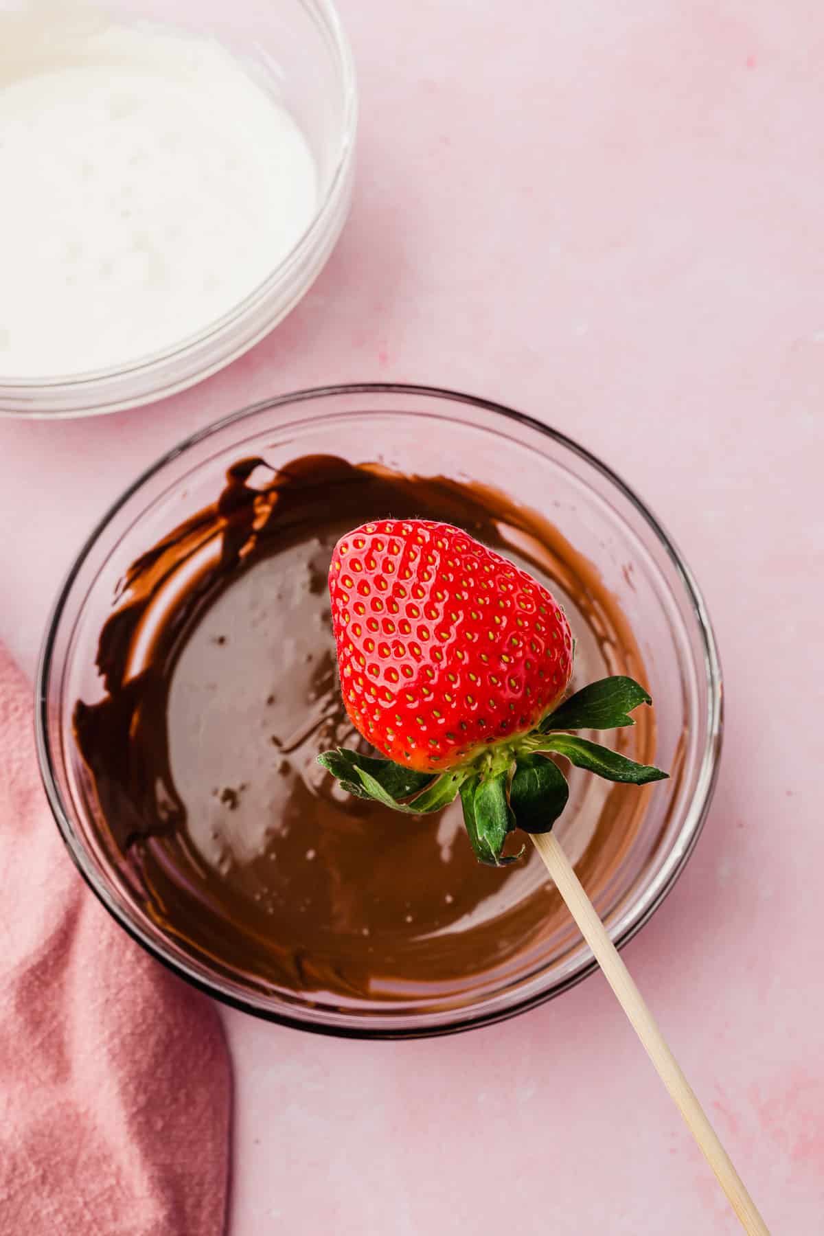 A fresh strawberry on a wooden skewer being held over a bowl of melted dark chocolate before dipping.