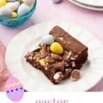 A single Easter mini egg brownie on a dessert plate with a bowl of mini chocolate eggs in the background.