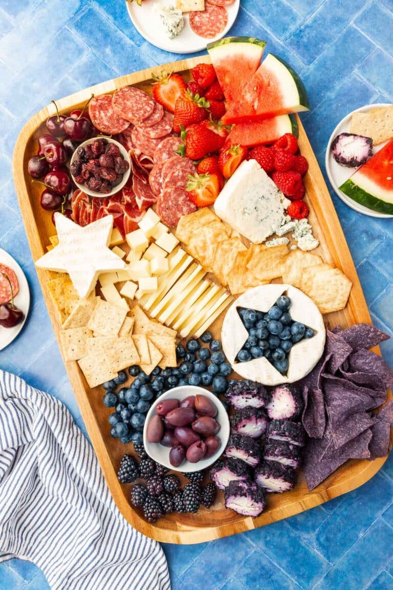 An overhead view of a 4th of July charcuterie board on a blue table that is filled with red, white and blue ingredients like meat, cheeses, crackers and fresh fruits.