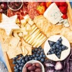 An overhead view of a July 4th charcuterie board including meats, cheeses, crackers, fresh fruit and a round of brie with a star cut out of it and filled with blueberries.