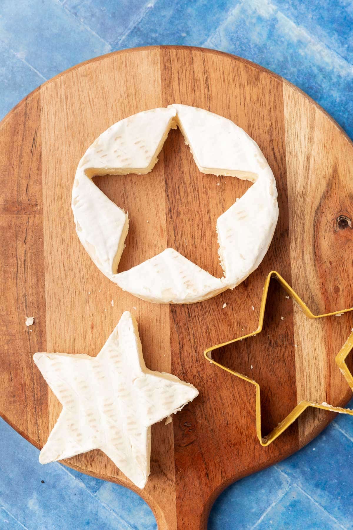 A small wood cutting board with a brie round that has a star shape cut out of it with a star cookie cutter and the center of the brie next to it.