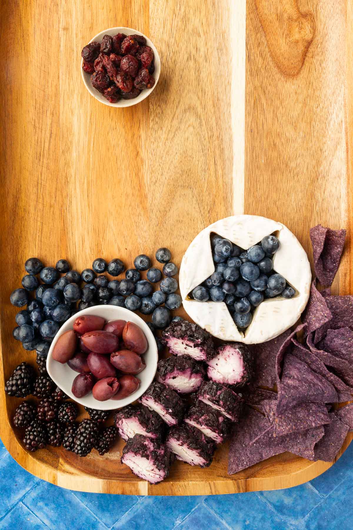 A wood charcuterie board with a round brie with a star cut out of it and filled with blueberries, a blueberry goat cheese log, blue corn tortilla chips, blackberries, a bowl of kalamata olives, and a bowl of dried cranberries.