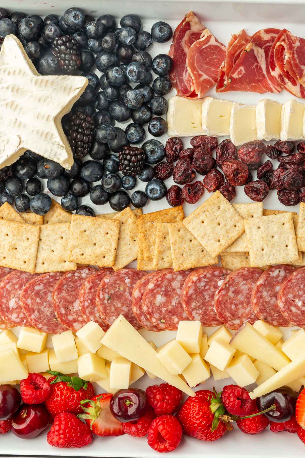A close up of an American flag charcuterie board including blueberries, blackberries, a brie star, capicola, dried cranberries, salami, crackers, cheddar cheese, cherries, raspberries and strawberries.