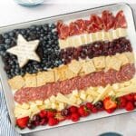 An overhead view of a 4th of July charcuterie board in the shape of an American flag, including a brie star, blueberries, dried cranberries, meats, cheddar cheese, crackers, strawberries, raspberries and cherries.