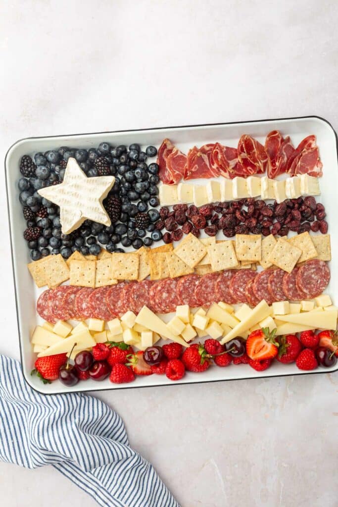A white sheet pan with blueberries, blackberries, cured meats, cheese, crackers, raspberries, strawberries, and cherries assembled to look like an American flag.