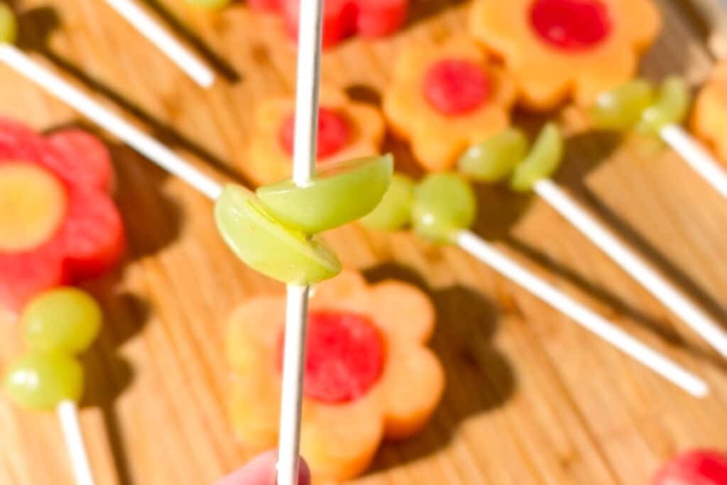 A close-up of green grape halves being skewered onto a white stick as part of a flower fruit kabob.