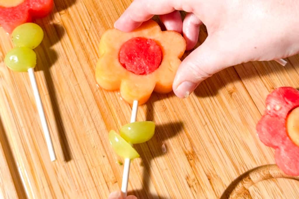A hand placing a cantaloupe flower with a watermelon center onto a fruit kabob skewer topped with grapes.