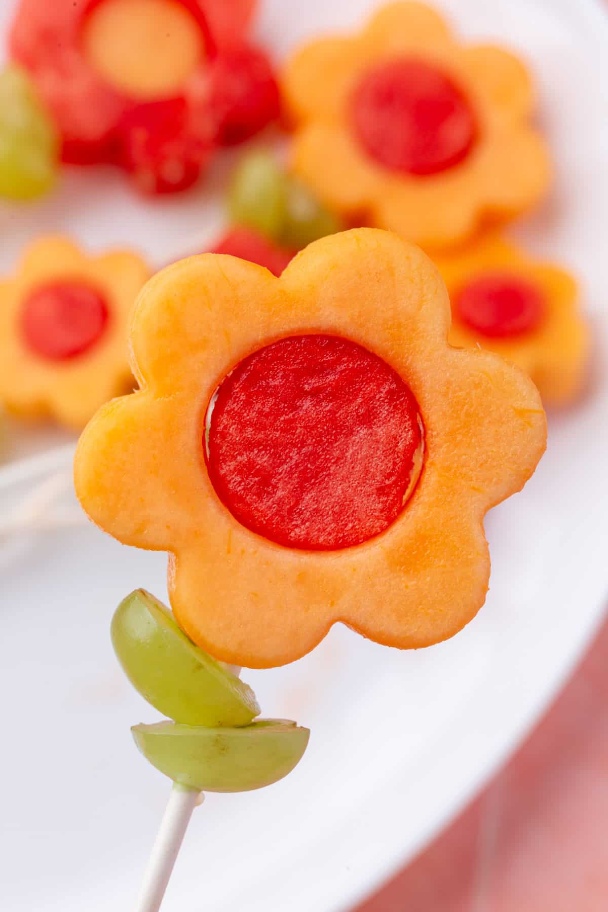A close-up of a flower-shaped cantaloupe slice with a watermelon center and green grape halves on a skewer.